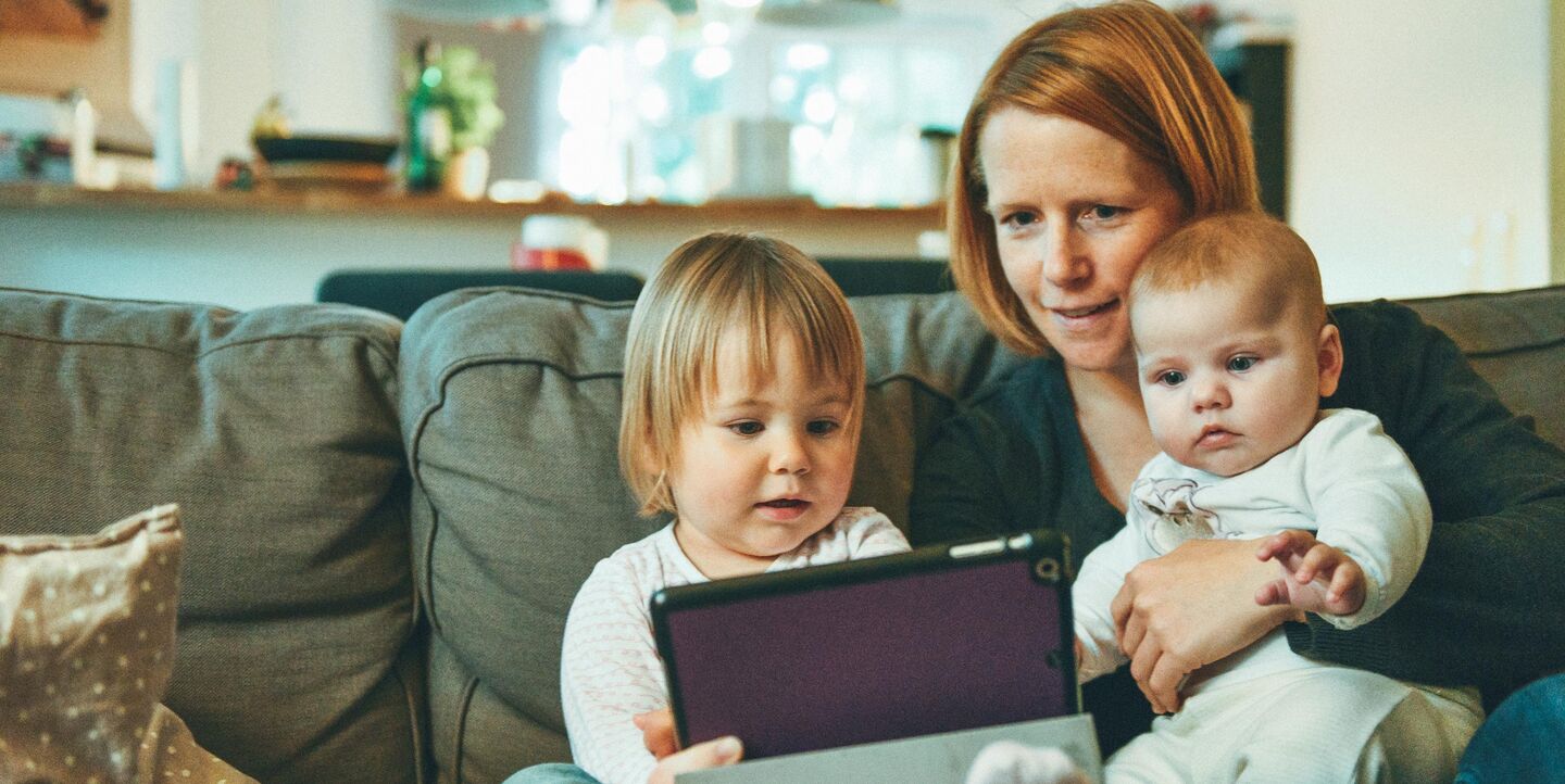 Eine Mutter und zwei Kindersitzen in ihrem Wohnzimmer auf ihrer Couch und schauen auf den Bildschirm ihres Ipads.